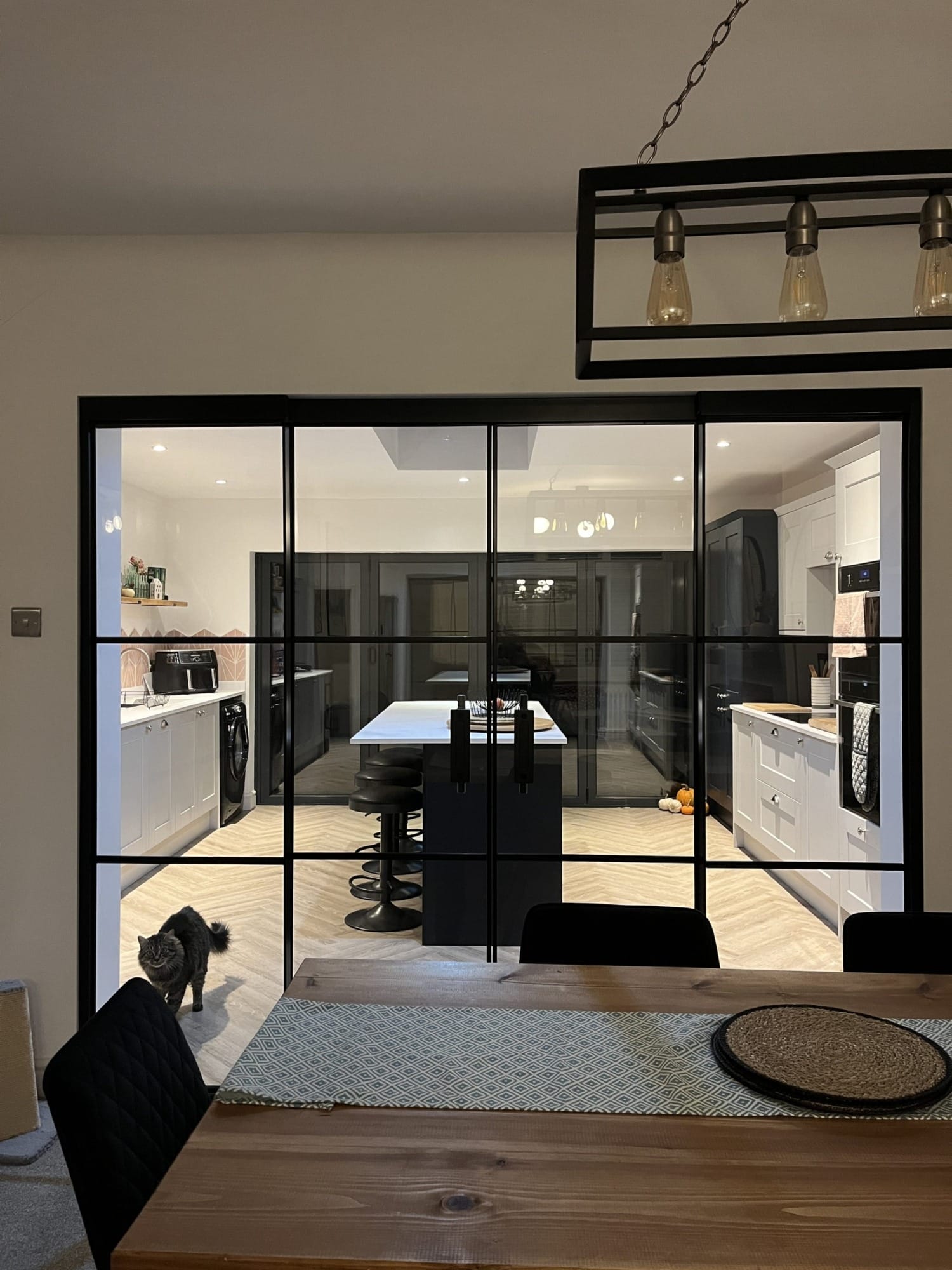 A set of black-framed partition doors made of glass, dividing a dining area from a modern kitchen. The doors feature a grid-like design with thin, black metal frames. In the foreground is a wooden dining table with a patterned runner and round woven placemats. The kitchen behind the doors is brightly lit and features a central island with black barstools, white countertops, and sleek cabinetry in white and dark tones. A gray fluffy cat is walking through the doorway on the left side. The floor has a light herringbone wood pattern, and there are small decorative items visible, including pumpkins on the kitchen floor. A black rectangular pendant light fixture with exposed bulbs hangs from the ceiling above the table.