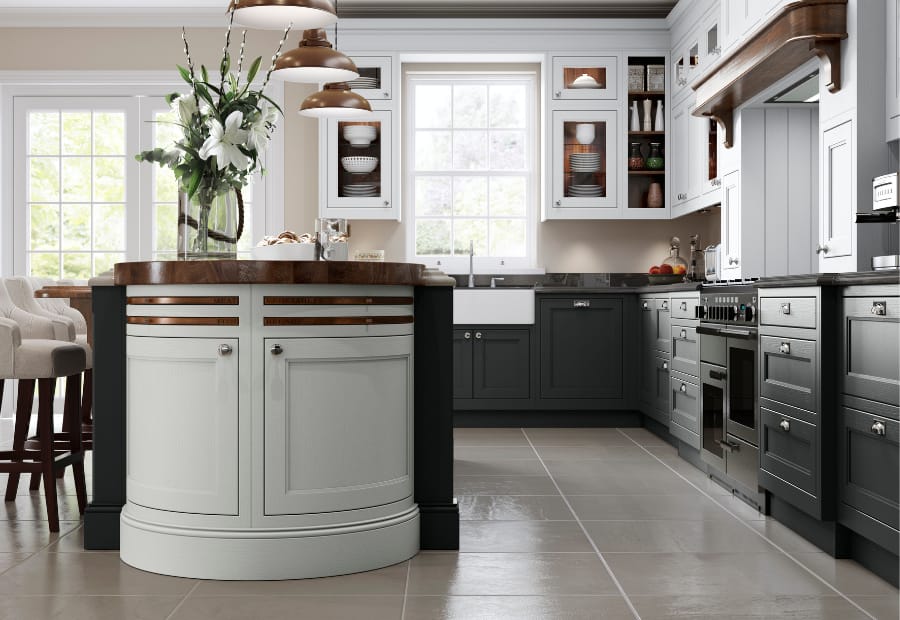 A sophisticated kitchen design featuring a two-tone colour scheme of soft shell and gunmetal grey cabinetry. The centrepiece is a curved kitchen island with a rich wooden worktop and integrated storage, complemented by elegant copper pendant lighting above. The perimeter cabinets offer ample storage and are paired with sleek black granite countertops. A large farmhouse sink sits beneath a bright window, flooding the space with natural light. Open shelving and glass-fronted upper cabinets add charm and practicality, while the tiled flooring completes the room with a polished and timeless look. Perfectly blending classic and contemporary elements, this kitchen exudes elegance and functionality.