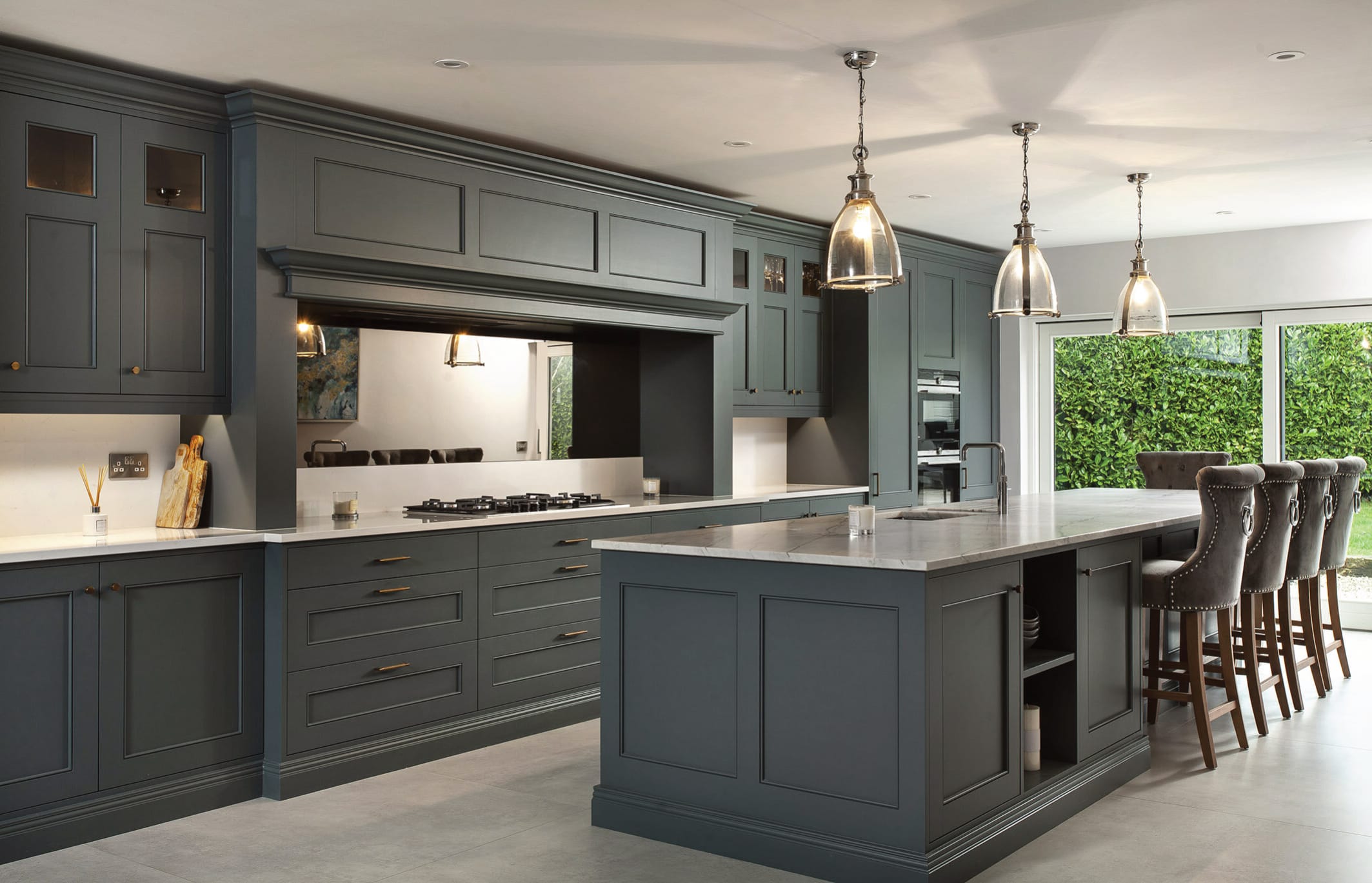 A luxurious and bespoke kitchen featuring a sophisticated in-frame Shaker design in a rich grey-green hue. The cabinetry is complemented by elegant brass handles, a central island with a marble-effect worktop, and integrated shelving for added functionality. Pendant lights with glass shades hang above the island, adding a touch of timeless charm. The kitchen includes a gas hob with a concealed extractor fan, double ovens, and a spacious breakfast bar with studded upholstered bar stools. Large sliding glass doors in the background bring in natural light and offer a view of a lush, green garden. The space exudes both functionality and high-end design.